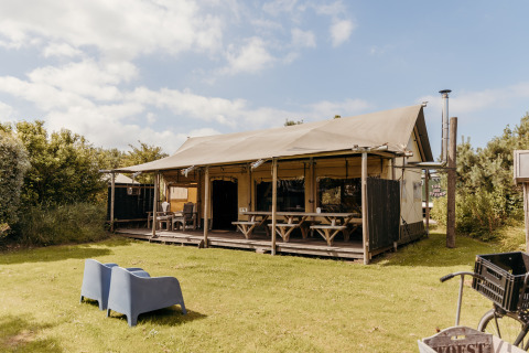 La tente safari Wood Lodge avec terrasse abritée, bancs et chaises sur une pelouse verte sous ciel bleu.