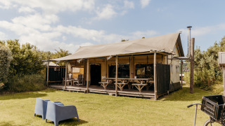 La tente safari Wood Lodge avec terrasse abritée, bancs et chaises sur une pelouse verte sous ciel bleu.