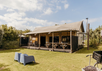 La tenda safari Wood Lodge con veranda coperta, tavoli da picnic e sedie su un prato verde e cielo sereno.