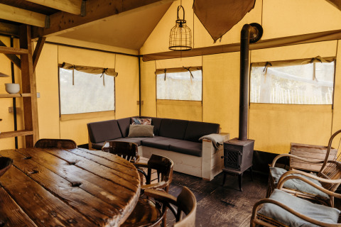 Vista interior de una tienda safari Wood Lodge en Duynpark Het Zwanenwater, con sofá, estufa y mesa comedor.