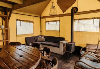 Vista interior de una tienda safari Wood Lodge en Duynpark Het Zwanenwater, con sofá, estufa y mesa comedor.