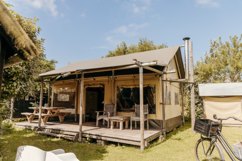 Tienda safari Beach Lodge con porche, asientos al aire libre, mesas de picnic y bicicleta en entorno verde.
