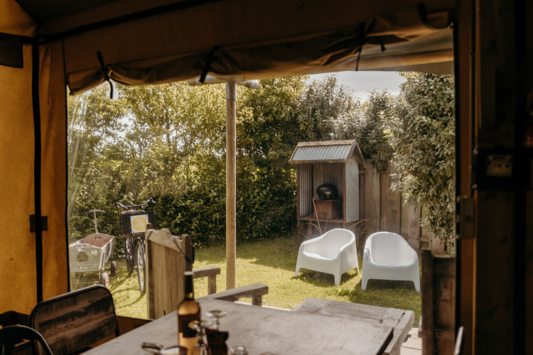 Uitzicht vanuit een safaritent op tuin met witte stoelen, barbecue en fietsen bij Duynpark Het Zwanenwater.