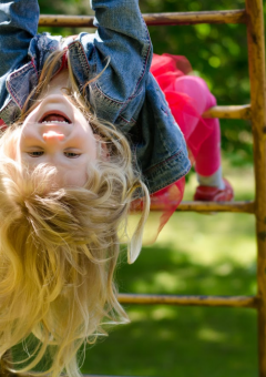 Una bambina sorridente appesa a testa in giù su una struttura da gioco in un parco glamping immerso nel verde.