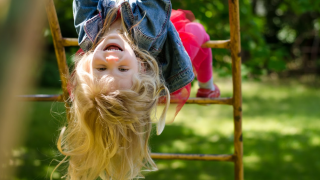 Una bambina sorridente appesa a testa in giù su una struttura da gioco in un parco glamping immerso nel verde.