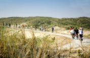 Ciclistas recorren un sendero arenoso y rodeado de pasto cerca de Hollum, Frisia, Países Bajos, bajo cielo azul.