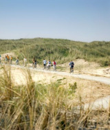 Cykelryttere på en sti omgivet af klitter og græs nær Hollum, Friesland, Nederlandene, under blå himmel.