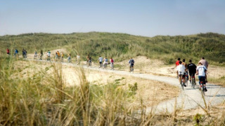 Ciclistas recorren un sendero arenoso y rodeado de pasto cerca de Hollum, Frisia, Países Bajos, bajo cielo azul.