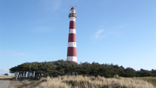 Faro de franjas rojas y blancas cerca de Hollum, Frisia, Países Bajos, rodeado de dunas y pinos bajo un cielo azul claro.