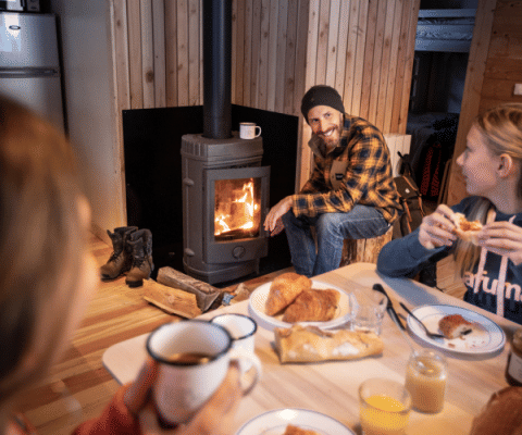 Family enjoys breakfast around a wood stove in a cabin at Les Chalets Huttopia Champagny en Vanoise.