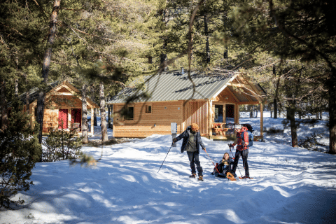 Familie beim Wintersport vor gemütlichen Holzhütten in verschneitem Wald bei Huttopia Champagny en Vanoise.