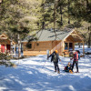 Family enjoys winter activities outside wooden cabins in snowy forest at Les Chalets Huttopia Champagny en Vanoise.