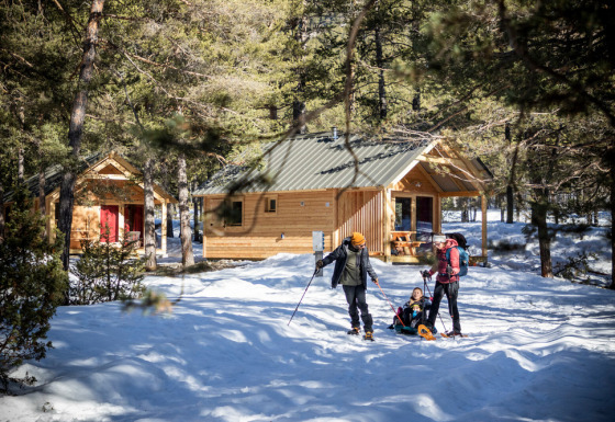 Familia disfruta actividades de invierno frente a cabañas en bosque nevado en Les Chalets Huttopia Champagny en Vanoise.