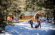 Familie beim Wintersport vor gemütlichen Holzhütten in verschneitem Wald bei Huttopia Champagny en Vanoise.