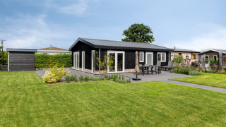 Moderne lodge ved navn Natuurhoeve med sort facade, stor terrasse og grøn have under blå himmel.