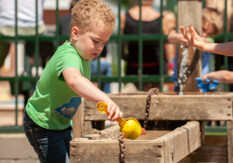 Un bambino con maglietta verde gioca con l'acqua e una paletta gialla al Camping Goorzicht in Olanda.