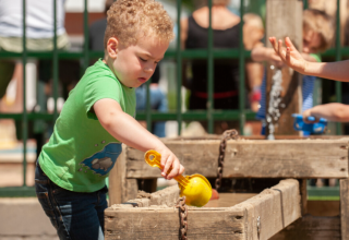 Un bambino con maglietta verde gioca con l'acqua e una paletta gialla al Camping Goorzicht in Olanda.