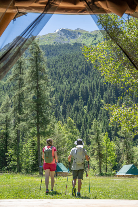 Dos excursionistas salen de una tienda glamping en Huttopia Vanoise, rodeados de montañas y bosques verdes.