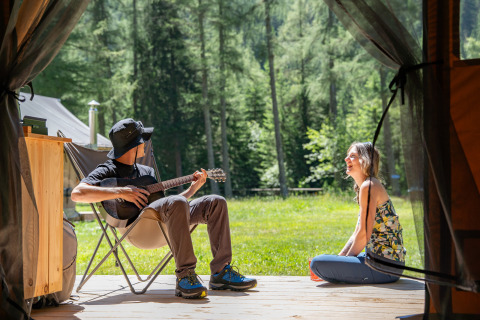 To personer hygger sig foran et glamping-telt i Huttopia Vanoise i Auvergne-Rhône-Alpes, en spiller guitar.