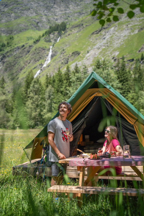 Deux personnes profitent du glamping devant une tente dans les montagnes vertes d’Huttopia Vanoise – Champagny.