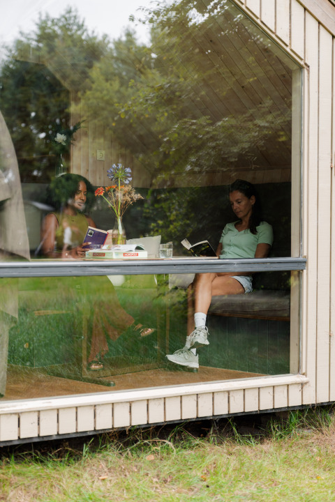 Two women reading books at a table in a tiny house with large windows at RØSTIG Dorst in the Netherlands.
