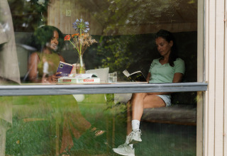 Two women reading books at a table in a tiny house with large windows at RØSTIG Dorst in the Netherlands.