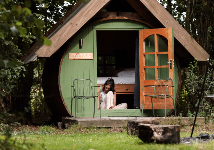 Una mujer se sienta en la entrada de una cabina verde SOLO TØN en el bosque, rodeada de naturaleza y árboles.