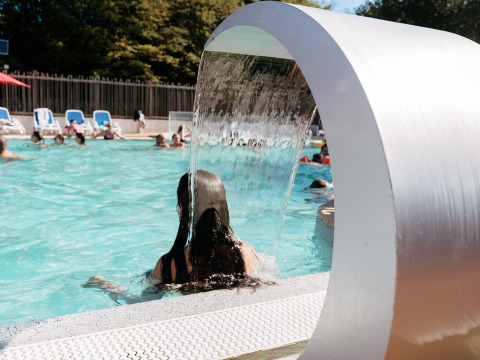 Person standing under a water feature in an outdoor pool at Flower Camping Le Kerarno, Brittany, France.