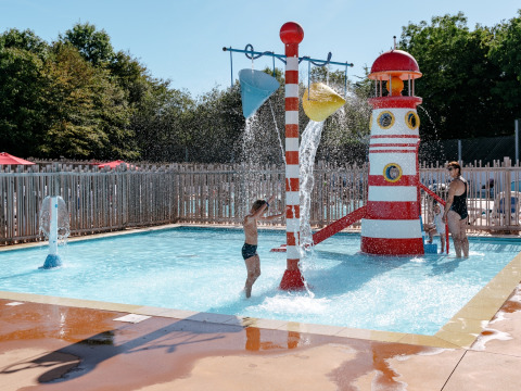 Children play in a shallow pool with a water playground and slide at Flower Camping Le Kerarno, France.