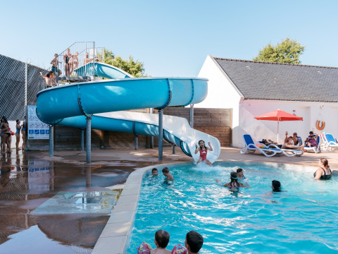 Familias disfrutan junto a la piscina y un gran tobogán azul en Flower Camping Le Kerarno en Bretaña, Francia.