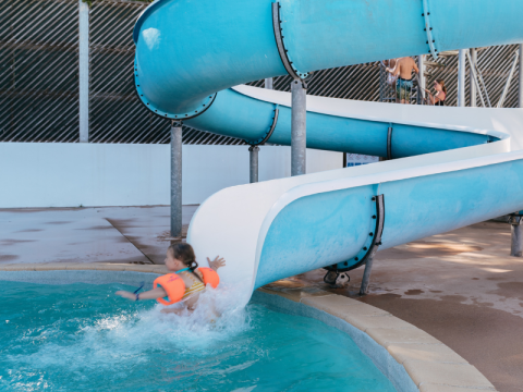 Et barn med orange badevinger lander i poolen fra en blå rutsjebane i Flower Camping Le Kerarno, Bretagne.