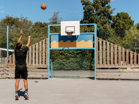 A man shoots a basketball into a hoop on an outdoor court at Flower Camping Le Kerarno in Brittany, France.