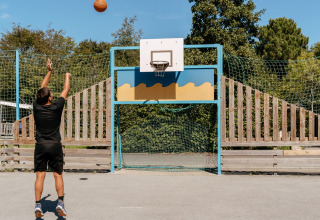 A man shoots a basketball into a hoop on an outdoor court at Flower Camping Le Kerarno in Brittany, France.