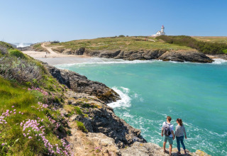 Couple stands on rocks overlooking turquoise bay, sandy beach, wildflowers, and lighthouse, Brittany, France.