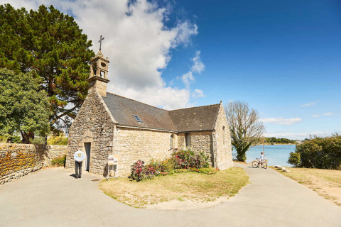 Piccola cappella di pietra a Saint-Philibert, Bretagna, Francia, con ciclisti e fiori vicino a un lago.