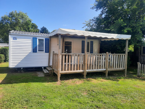 Mobile home Stacaravan Confort 28m2 with wooden porch, canopy, and blue shutters, on grassy lawn.