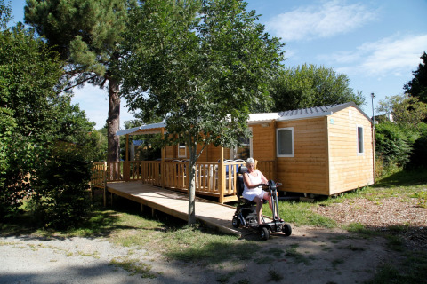 Mujer en scooter eléctrico frente a la casa móvil Comfort Stacaravan PMR 29m2 con rampa en entorno natural.