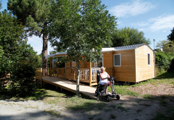Mujer en scooter eléctrico frente a la casa móvil Comfort Stacaravan PMR 29m2 con rampa en entorno natural.