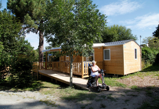 Femme sur scooter électrique devant la mobil-home Comfort Stacaravan PMR 29m2 avec rampe en plein air.