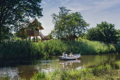Familie paddelt im Kanu auf Fluss neben Baumhaus in Holiday Park Mölke, Overijssel, Niederlande, Sommertag.