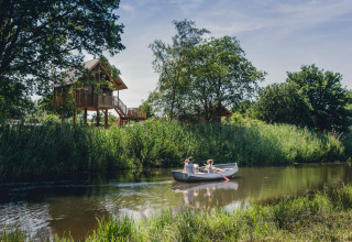 Familie paddelt im Kanu auf Fluss neben Baumhaus in Holiday Park Mölke, Overijssel, Niederlande, Sommertag.