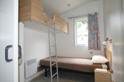 Modern mobile home bedroom with bunk bed, single bed, window, and colorful curtain at Camping Le Kerarno.