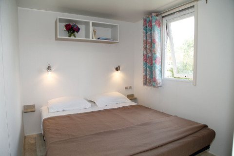 Camera da letto in una casa mobile con letto matrimoniale, finestra aperta, tende colorate e mensola.