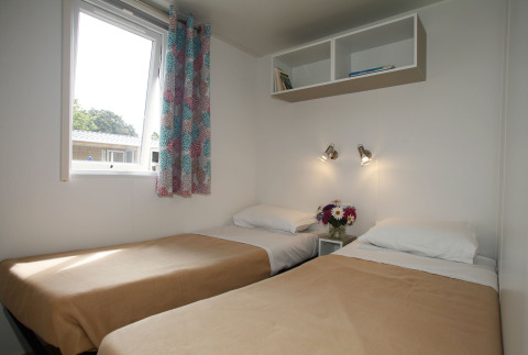 Chambre lumineuse en mobil-home avec deux lits simples, rideaux colorés et fleurs sur la table de nuit.