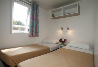 Chambre lumineuse en mobil-home avec deux lits simples, rideaux colorés et fleurs sur la table de nuit.