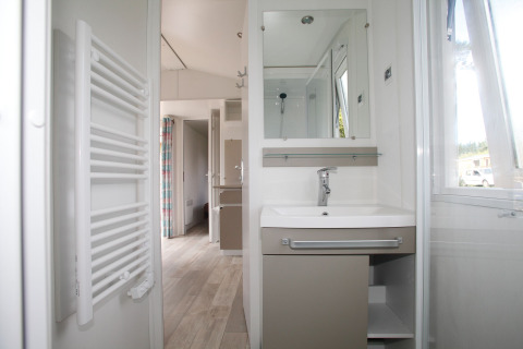 Salle de bain dans une maison mobile avec lavabo, miroir, radiateur porte-serviettes et fenêtre.