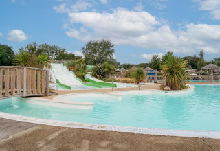 Buitenzwembad met waterglijbanen, palmbomen en rieten parasols bij Flower Camping Le Vorlen in Bretagne.