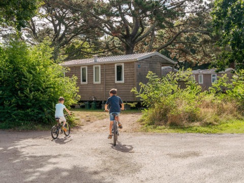 To børn cykler mod et træhus omgivet af grønt ved Flower Camping Le Vorlen-feriestedet i Bretagne, Frankrig.