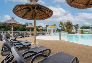 Entspannungsbereich mit Sonnenliegen und Strohdächern am Pool im Flower Camping Le Vorlen, Bretagne, Frankreich.