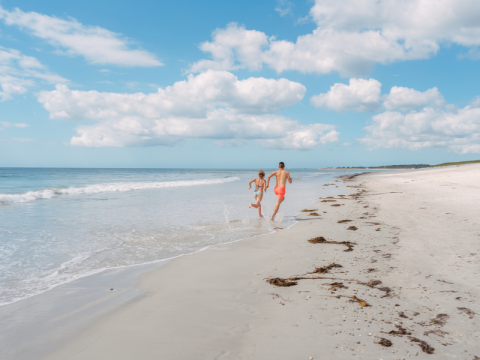 Two people run along a sunny sandy beach by the sea with blue skies at Flower Camping Le Vorlen, Brittany.